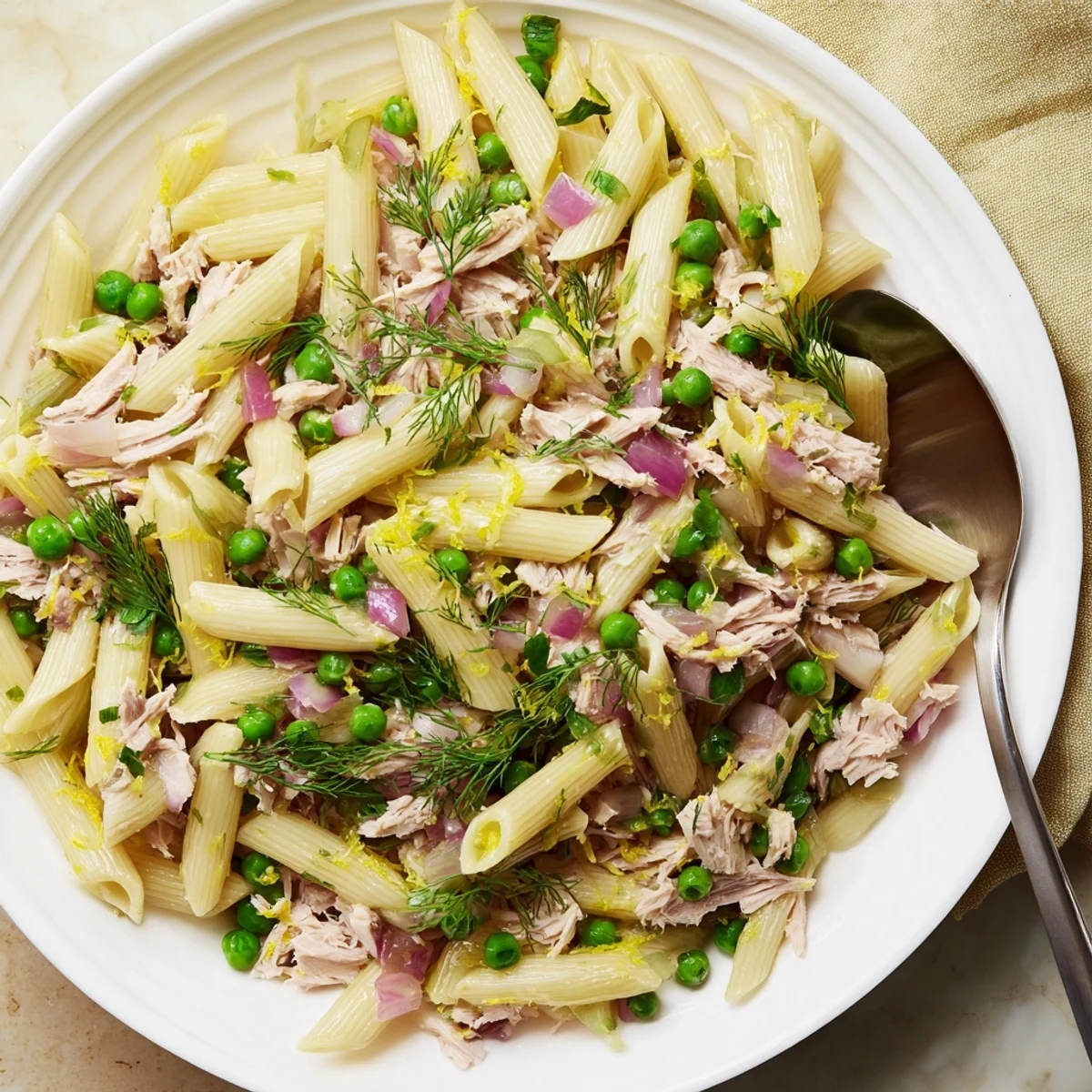 Bright Lemony Tuna Pasta Salad served in a white bowl, featuring golden pasta, tender tuna flakes, sweet green peas, and fresh dill garnish, drizzled with olive oil.  